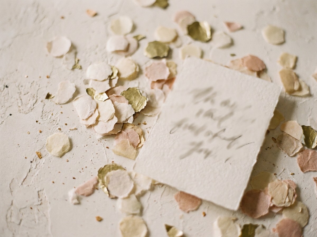 Scattered paper confetti in cream, blush and gold across a pale surface